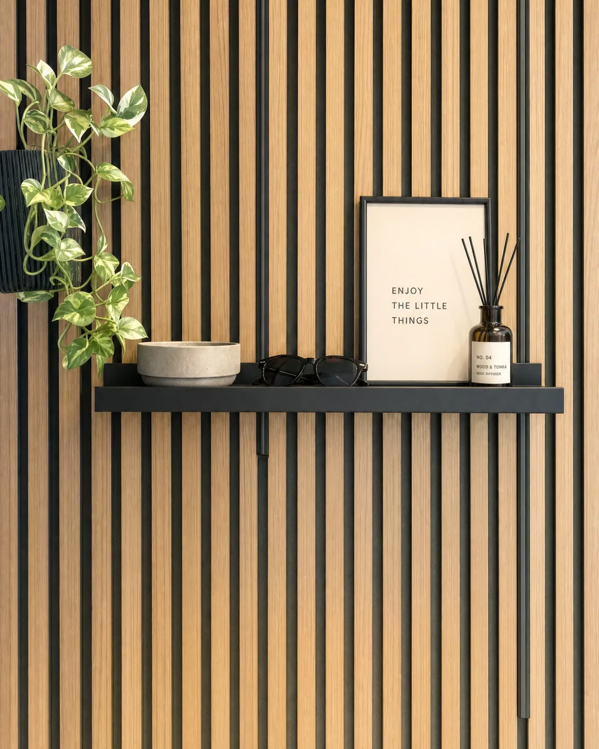 A black picture ledge with a framed print, a bowl, sunglasses and a reed diffuser, hung on a wood-slat acoustic panel by slim black brackets, beside a ribbed planter with a pothos