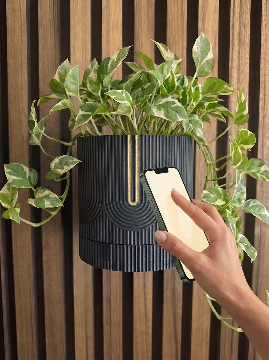 A hand holds a phone against a matte black ribbed wall planter with a thin gold line; the pot holds a trailing pothos on a walnut slat wall